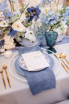 a blue and white table setting with flowers in vases, napkins and goldware