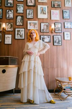 a woman standing in front of a wall with pictures on it and wearing a white dress