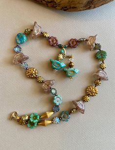 Here is a sweet shimmery bouquet of a necklace with golden AB Czech glass dahlias and soft rose and gold birds, two more aqua and gold birds suspended below.  The birds are 22mm, a translucent blush of rose with gold highlights. They are simple and charming. The dahlias, in contrast, are splashy and flashy-shimmery gold on one side and AB pink on the other.  The larger dahlias are 14mm and the little ones about half that. There are also a few Czech Glass daisies and leaves, all in shades of aqua. The spacers are small coppery beads and two vintage brass bicones are accent beads, their finely etched surface giving them a pretty shimmer. Two aqua birds dangle below the beaded necklace, one on a few links of chain for more length.  The necklace is 17" long and the bird dangles add another two Gold Flower-shaped Glass Jewelry, Gold Glass Flower Jewelry, Gold Glass Flower Shaped Jewelry, Gold Czech Glass Flower Jewelry, Gold Czech Glass Flower Shaped Jewelry, Whimsical Gold Jewelry With Czech Glass, Whimsical Gold Czech Glass Jewelry, Gold Beaded Necklaces With Flower Charm, Gold Beaded Necklace With Flower Charm As Gift