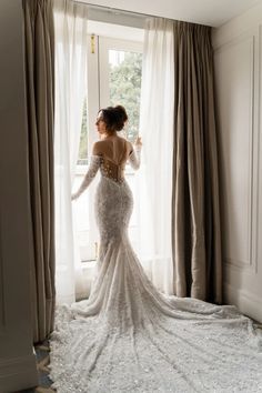 a woman standing in front of a window wearing a wedding dress with long sleeves and an open back