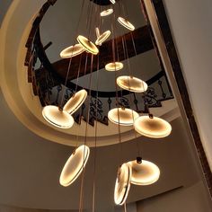 a circular chandelier with lights hanging from it's sides in front of a spiral staircase