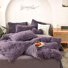 a bed with purple comforter and pillows in a room next to a plant on the floor