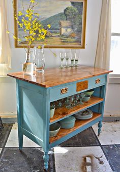 a blue table with vases and glasses on it in front of a framed painting