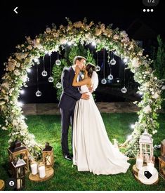 a bride and groom kissing in front of an archway decorated with white flowers at night