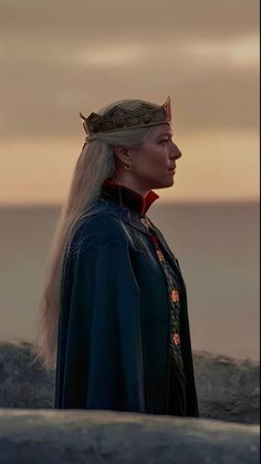 a woman with long blonde hair and a crown on her head standing in front of the ocean