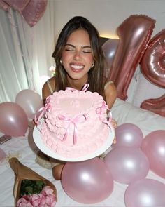 a woman holding a pink birthday cake in front of balloons and flowers on a bed