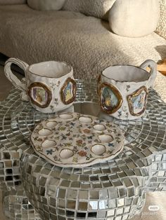 two coffee mugs sitting on top of a glass table next to a mirror plate