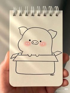 a hand holding a notebook with a drawing of a pig in a box on it