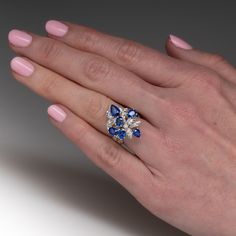 This captivating cluster style ring is accented with four (4), prong set, oval mixed cut natural sapphires and two (2), prong set, pear mixed cut natural sapphire. The sapphires are bordered with two (2), prong set, round brilliant cut diamonds and fourteen (14), bead set, round brilliant/single cut diamonds, bordered with milgrain edging. Engraved details accent the shoulders of the shank. The ring measures 22.6mm at the top, rises 9.8mm above the finger, tapering to 3.8mm wide and 1.3mm thick at the base of the shank. This ring is currently as size 6.5. Cluster Sapphire Ring With Accent Stones, Cluster Sapphire Ring In Fine Jewelry Style, Pear-shaped Multi-stone Sapphire Ring For Anniversary, Marquise Cut Sapphire Cluster Ring With Gemstones, Marquise Sapphire Ring With Rose Cut Diamonds, Luxury Cluster Sapphire Ring Gia Certified, Brilliant Cut Sapphire Cluster Jewelry, Classic Gia Certified Sapphire Cluster Ring, Blue Sapphire Cluster Ring Fine Jewelry