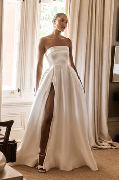 a woman standing in front of a window wearing a white dress with high slits