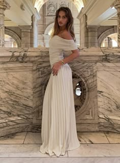 a woman standing in front of a marble wall wearing a white dress and gold jewelry