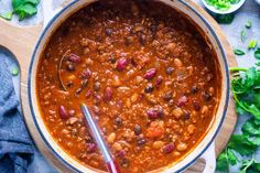 a pot filled with chili and beans on top of a wooden cutting board next to green vegetables