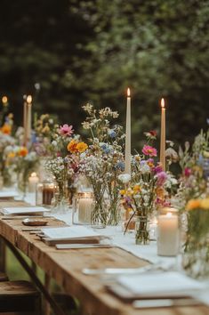 a long wooden table topped with lots of flowers and candles next to tall vases filled with greenery