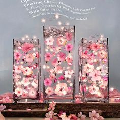 three glass vases with pink flowers in them on a wooden table next to some lights