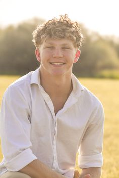 a young man sitting on the ground in front of a field with grass and trees