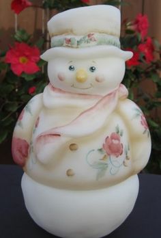 a ceramic snowman with a hat and scarf on it's head sitting in front of some flowers