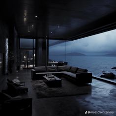 a living room filled with lots of furniture next to the ocean at night, in front of a large window