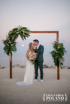 a bride and groom kissing under an arch with palm trees on the beach at sunset