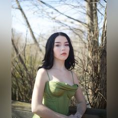 a woman in a green dress sitting on a wooden bench next to some bare trees