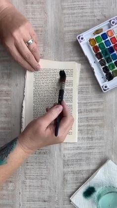 a person holding a paintbrush over an open book with watercolors on it