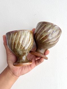 a hand holding two ceramic cups in front of a white wall