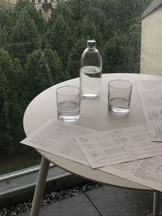 two glasses are sitting on a table next to some papers and a water bottle in front of a window