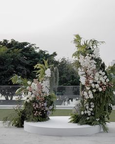 two white pedestals with flowers and greenery on them in the middle of a park