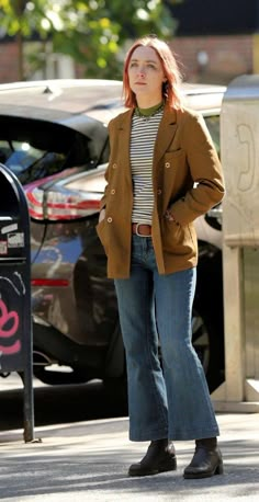 a woman with red hair is standing in the street wearing jeans and a brown jacket