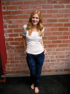 a woman standing in front of a brick wall with her hands in her pockets and smiling