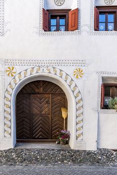 a white building with red shutters and two brown doors on each side, surrounded by stones