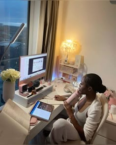 a woman sitting at a desk with a laptop and computer monitor on it, looking out the window