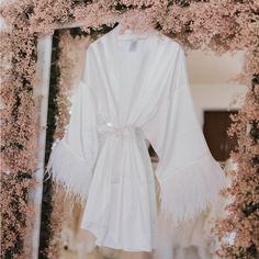 a white robe hanging in front of a mirror with pink flowers on the wall behind it