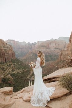 a woman in a wedding dress standing on top of a cliff with mountains behind her