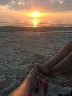 two people sitting on the beach at sunset with their feet up in the sand,