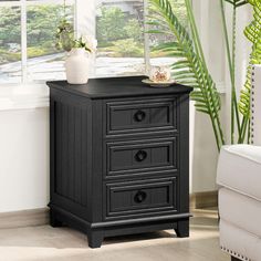 a black cabinet with three drawers in front of a white chair and potted plant