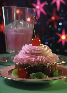 a cupcake with pink frosting and cherries sits on a plate next to a glass of milkshake