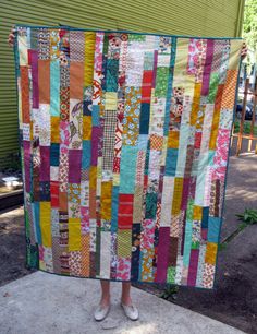 a woman holding up a colorful quilt on the side of a building with trees in the background