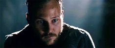 a man with a beard and blue eyes stares into the camera while standing in a dark room