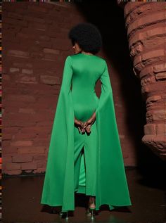 a woman in a green dress and cape standing next to a brick wall with her hands on her hips