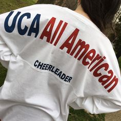 "UCA all American across the shoulders of this oversized jersey shirt and cheerleader in smaller letters under that line. This one has the British flag with name and year but it's your choice. Add matching bow! These run HUGE! The XS is about 42\" chest, S 44\" and so on. Order accordingly. Long sleeve, ring-spun cotton tshirt. Super soft . Please specify your lettering and colors allow 7-10 days processing." Varsity Tops For Cheerleading During Football Season, School Spirit T-shirt For Cheerleading In Fall, White Casual Top For Tailgating, Casual White Top For Tailgating, Cheerleading Fan Apparel Tops For Football Season, White Sports Fan College Top, Sporty Crew Neck Top For Tailgating, Team Spirit Crew Neck Top For Tailgating, Team Spirit Tops For Tailgating With Crew Neck