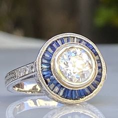 Details: Stunning vintage Art Deco style diamond & sapphire engagement ring. The band is platinum. This is an gorgeous ring with lovely engraving around the shoulders of the band, and has engraving details surrounding the diamond. This ring is stunning! Appraisal Reads: One old European cut diamond, measuring 7.20 x 7.20 x4.36mm an approx. weight 1.38ct. with faint blue fluorescences. Clarity H-I Cut Very Good Depth 60.6% Table 47.2% Six round brilliant cut diamonds measuring 1.5 x 1.5 x .90 Art Deco Gia Certified Sapphire Ring With Diamond, Art Deco Sapphire Ring With Gia Certified Diamond, Art Deco Gia Certified Sapphire And Diamond Ring, Art Deco Gia Certified Round Cut Sapphire Ring, Art Deco Sapphire Ring With Halo Setting, Vintage Silver Gia Certified Halo Ring, Vintage Silver Halo Ring Gia Certified, Art Deco Sapphire Ring With Platinum Center Stone, Vintage Gia Certified Sapphire Ring