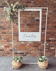 two planters with flowers are sitting in front of a brick wall and sign that says,