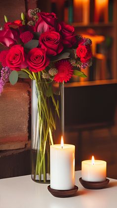 two white candles are next to a vase with red roses