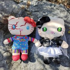 two hello kitty dolls sitting next to each other on a rock covered in leaves and grass