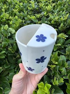 a hand holding up a blue and white vase in front of some green bushes with flowers on it