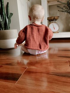 a toddler sitting on the floor looking at a potted plant with a clock in the background