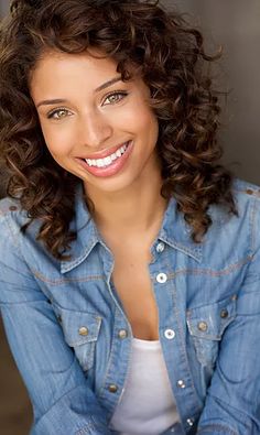 a woman with curly hair smiling at the camera and wearing a jean jacket over her shoulders