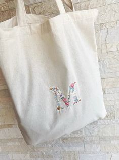 a white tote bag with embroidered letter m on it hanging from a brick wall