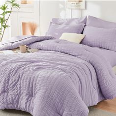 a bed with purple comforter and pillows on it in a room next to a potted plant