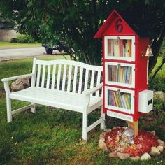 a white bench sitting next to a red book shelf on top of a grass covered field